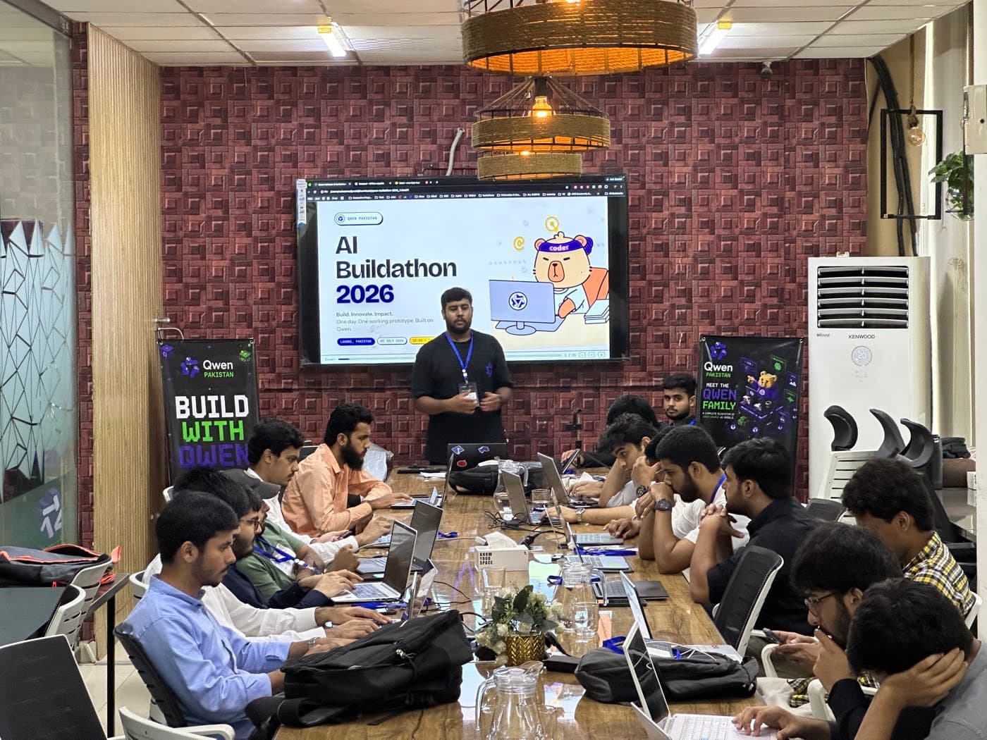 A long table of developers with laptops open at the Qwen Pakistan AI Buildathon, a slide reading AI Buildathon 2026 on the screen behind them